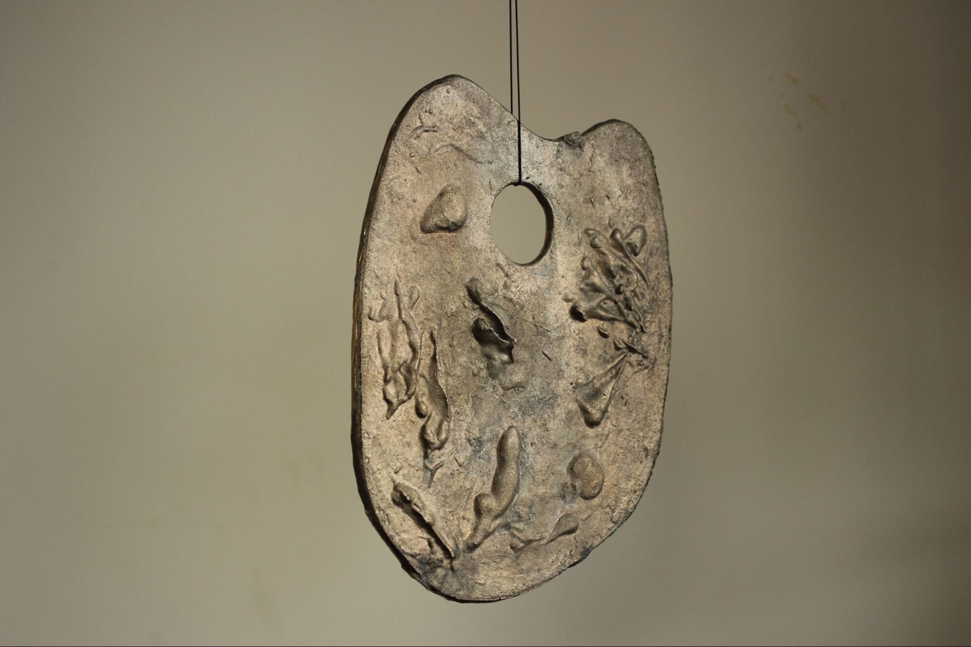 A bronze-coloured jewellery object with creased, organic shapes embossed into it, hanging from a string.