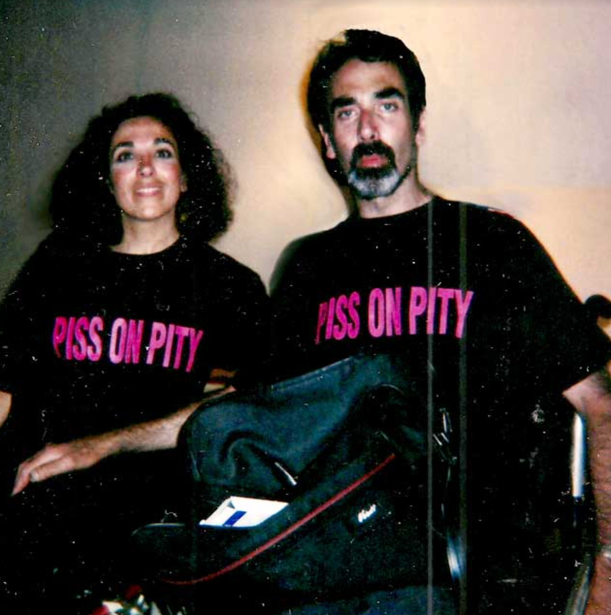 A man and a woman in wheelchairs, both wearing shirts that say Piss on Pity in bright pink capital letters.